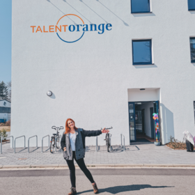 Eine junge rothaarige Frau steht vor dem Gebäude der TalentOrange Sprachschule. Das Gebäude ist weiß und trägt den Schriftzug TalentOrange in orange und blau. Die Frau zeigt einladend auf die geöffnete Tür. 
