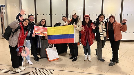 Acht junge OTA aus Kolumbien am Flughafen Hannover. Sie halten eine Kolumbienfahne und lächeln.
