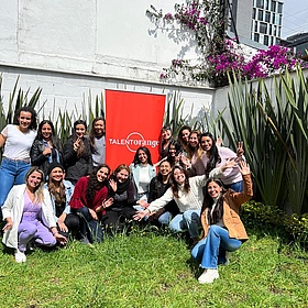 A group of language students in the garden of the TalentOrange language school in Bogota. They are sitting on a lawn. In the background is a white wall and a bush with purple flowers. Everyone is laughing happily.