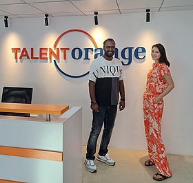Two TalentOrange employees are standing at the reception of the TalentOrange language school in Trivandrum. In the background you can see the lettering TalentOrange in blue and orange on the white wall.