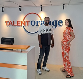 Zwei Mitarbeiter von TalentOrange stehen am Empfang der TalentOrange Sprachschule in Trivandrum. Im Hintergrund sieht man an der weißen Wand den Schriftzug TalentOrange in blau und orange. 